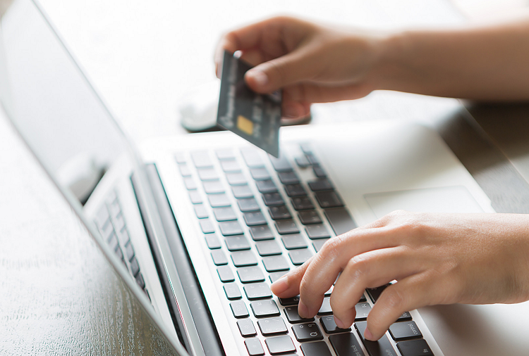 Da esquerda para a direto, um mac está sobre uma mesa de madeira, o aparelho está sendo utilizado por uma pessoa que segura um cartão de credito.