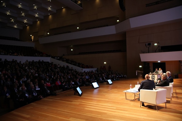 imagem de um auditório, da esquerda para a direta, plateia olha com atenção para 4 palestrantes sentados em poltronas sobre o palco.